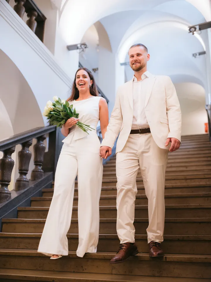 Brautpaar lacht auf der Treppe eines hellen Treppenhauses mit Gewölbedecke