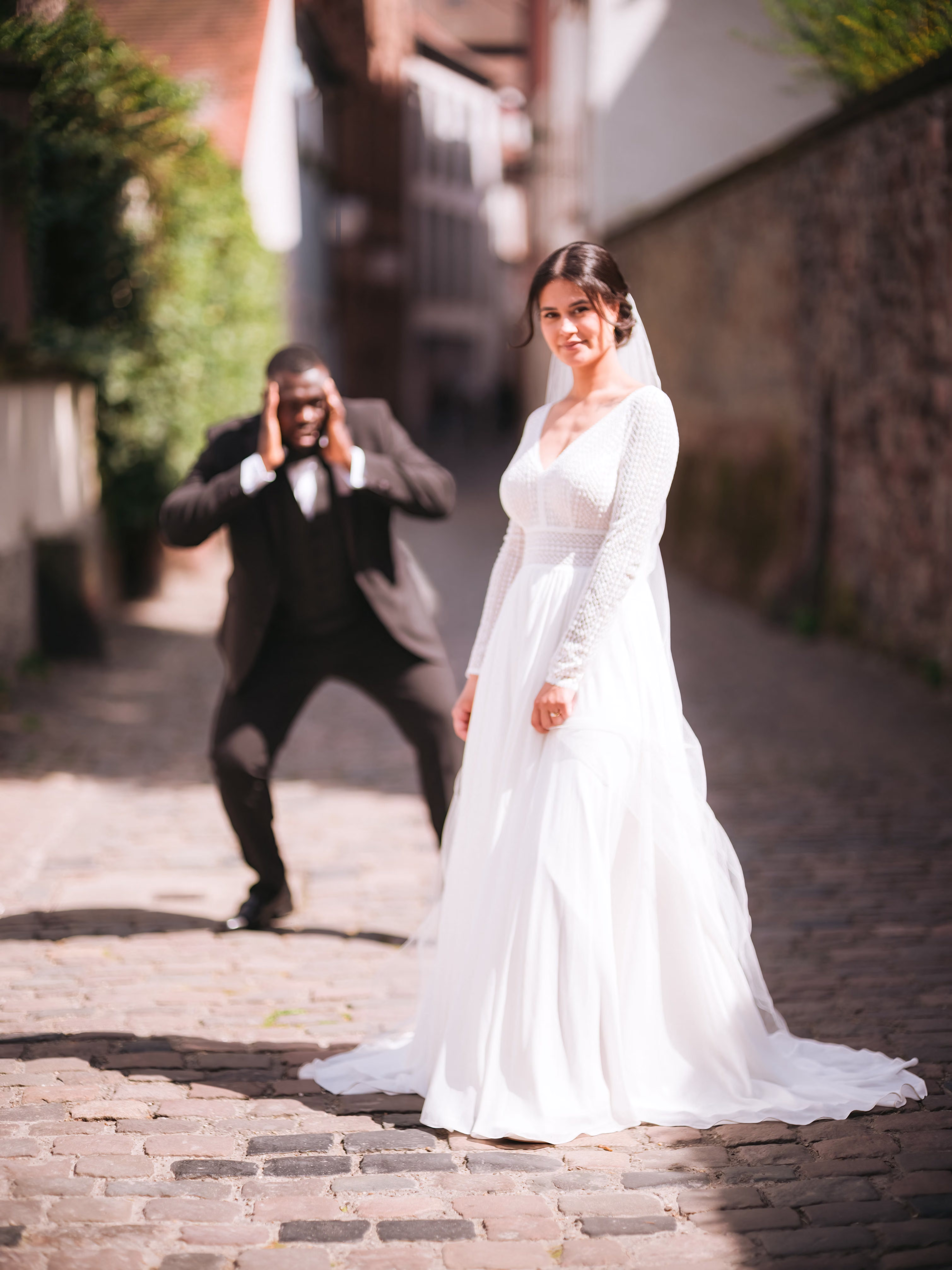 Braut im weißen Kleid in einer gepflasterten Altstadtgasse; eine Person im dunklen Anzug fotografiert sie – Bildstil Die Hochzeit meines besten Looks