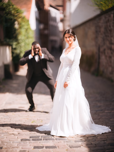 Braut im weißen Kleid in einer gepflasterten Altstadtgasse; eine Person im dunklen Anzug fotografiert sie – Bildstil Die Hochzeit meines besten Looks