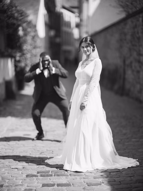 Braut im weißen Kleid in einer gepflasterten Altstadtgasse; eine Person im dunklen Anzug fotografiert sie – Bildstil My Big Fat Grey Wedding
