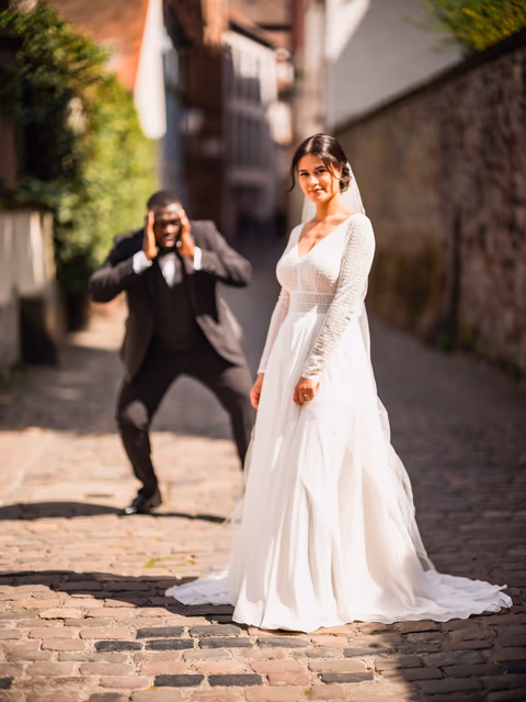 Braut im weißen Kleid in einer gepflasterten Altstadtgasse; eine Person im dunklen Anzug fotografiert sie – Bildstil 27 Hues