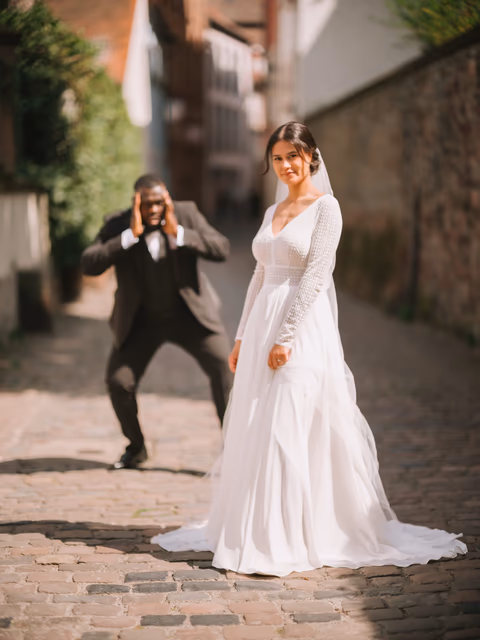 Braut im weißen Kleid in einer gepflasterten Altstadtgasse; eine Person im dunklen Anzug fotografiert sie – Bildstil Matte Mia!