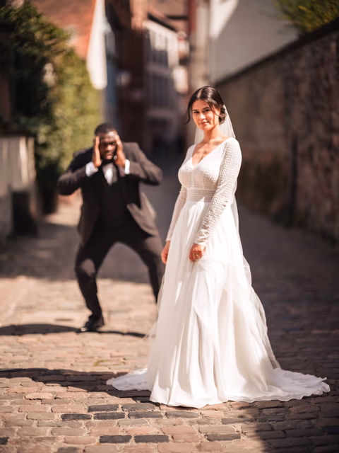 Braut im weißen Kleid in einer gepflasterten Altstadtgasse; eine Person im dunklen Anzug fotografiert sie – Bildstil Wedding Palette