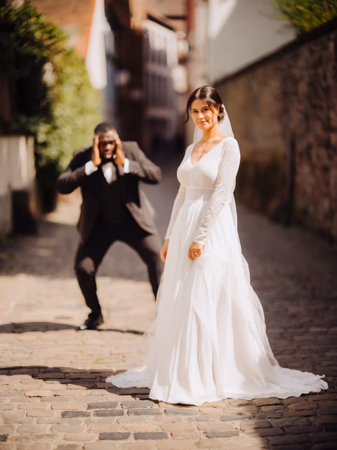Braut im weißen Kleid in einer gepflasterten Altstadtgasse; eine Person im dunklen Anzug fotografiert sie – Bildstil Bright Wars