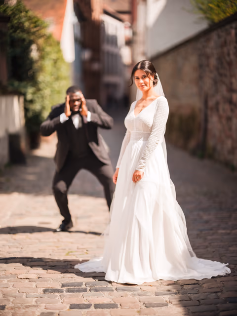 Braut im weißen Kleid in einer gepflasterten Altstadtgasse; eine Person im dunklen Anzug fotografiert sie – Bildstil Farbalarm