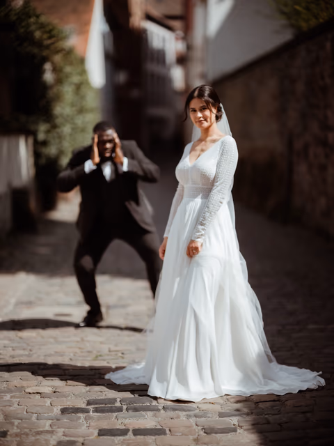 Braut im weißen Kleid in einer gepflasterten Altstadtgasse; eine Person im dunklen Anzug fotografiert sie – Bildstil Moodsoon Wedding