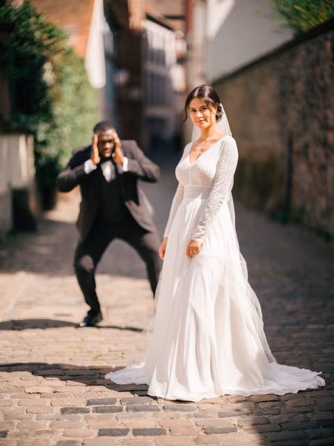 Braut im weißen Kleid in einer gepflasterten Altstadtgasse; eine Person im dunklen Anzug fotografiert sie – Bildstil Rachels Feinschliff
