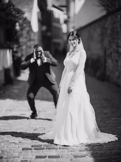 Braut im weißen Kleid in einer gepflasterten Altstadtgasse; eine Person im dunklen Anzug fotografiert sie – Bildstil Wedding Clashers
